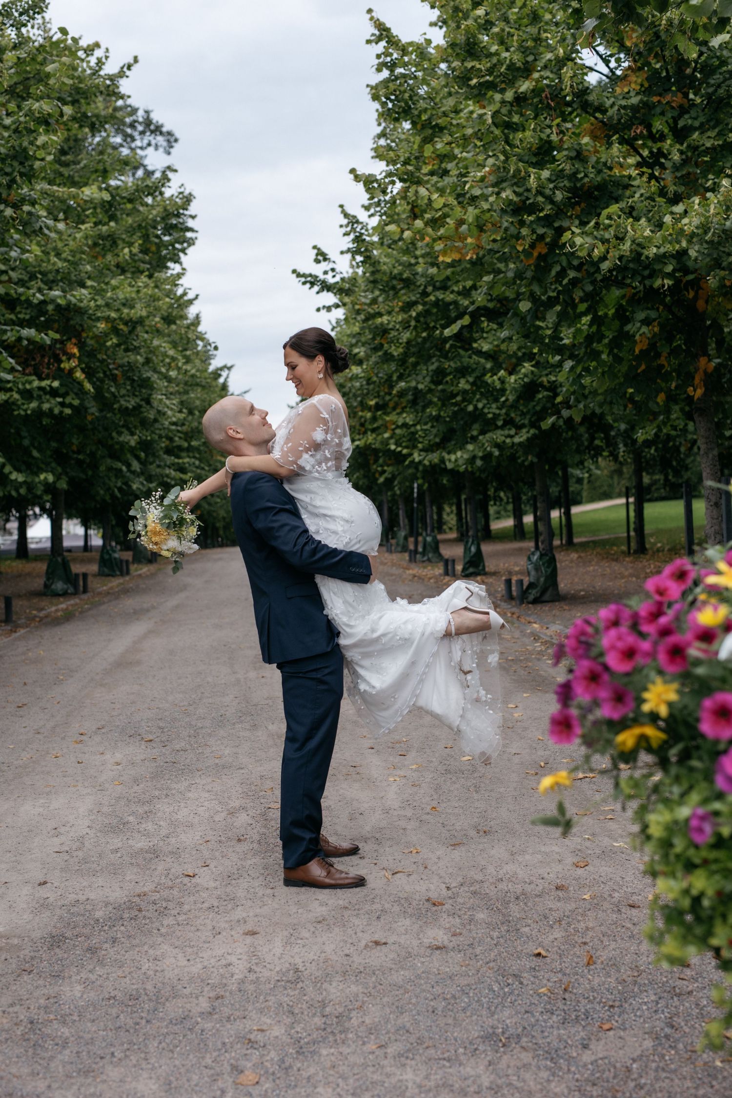 Groom lifts bride in white lace dress along tree-lined path, colorful flowers in foreground, outdoor wedding photo.
