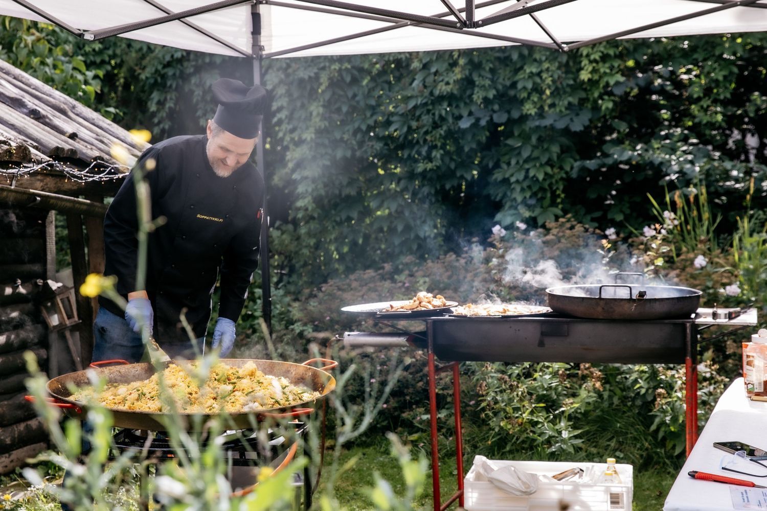 Chef cooking paella on large outdoor grills under a canopy at a garden catering event.