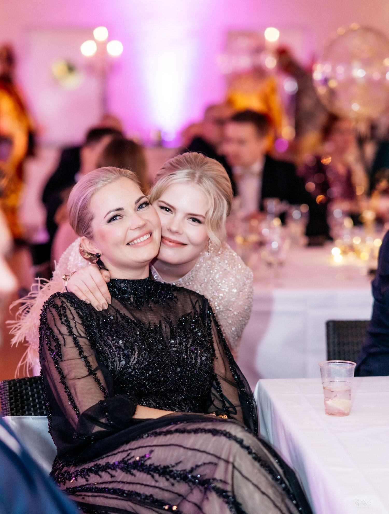 Two smiling blonde women in elegant sequined gowns embracing at a glamorous gala event.