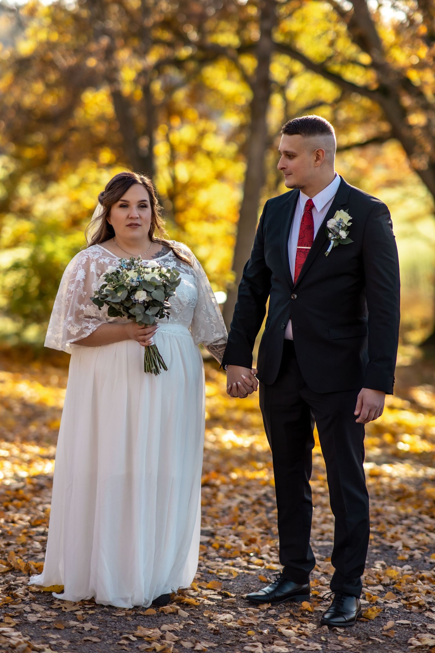 Bride and groom holding hands in autumn park, she in white gown with bouquet, he in black suit with red tie.