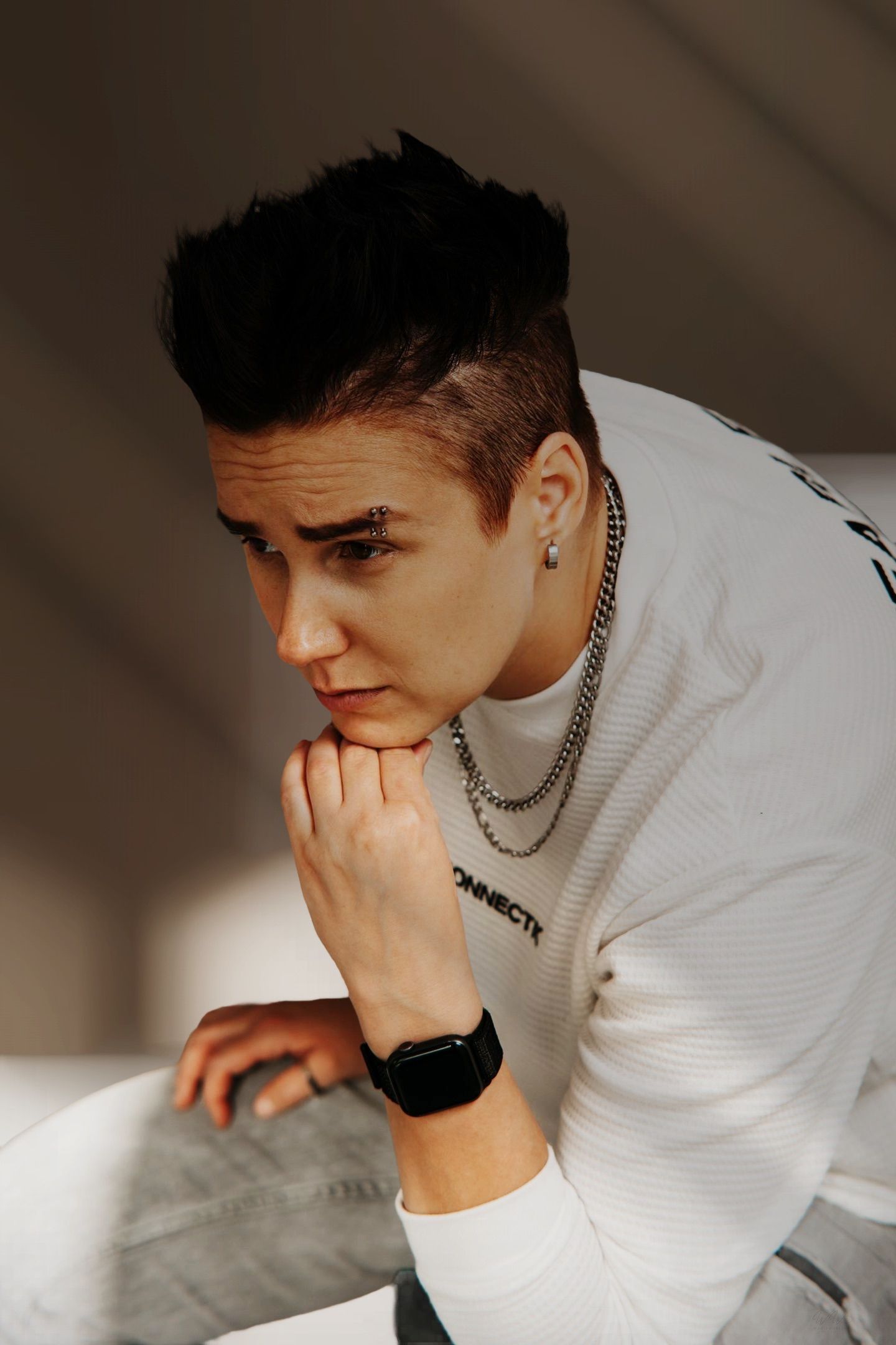 Person with short styled dark hair, white sweatshirt, layered silver chains and smartwatch, resting chin on hand in a thoughtful pose.