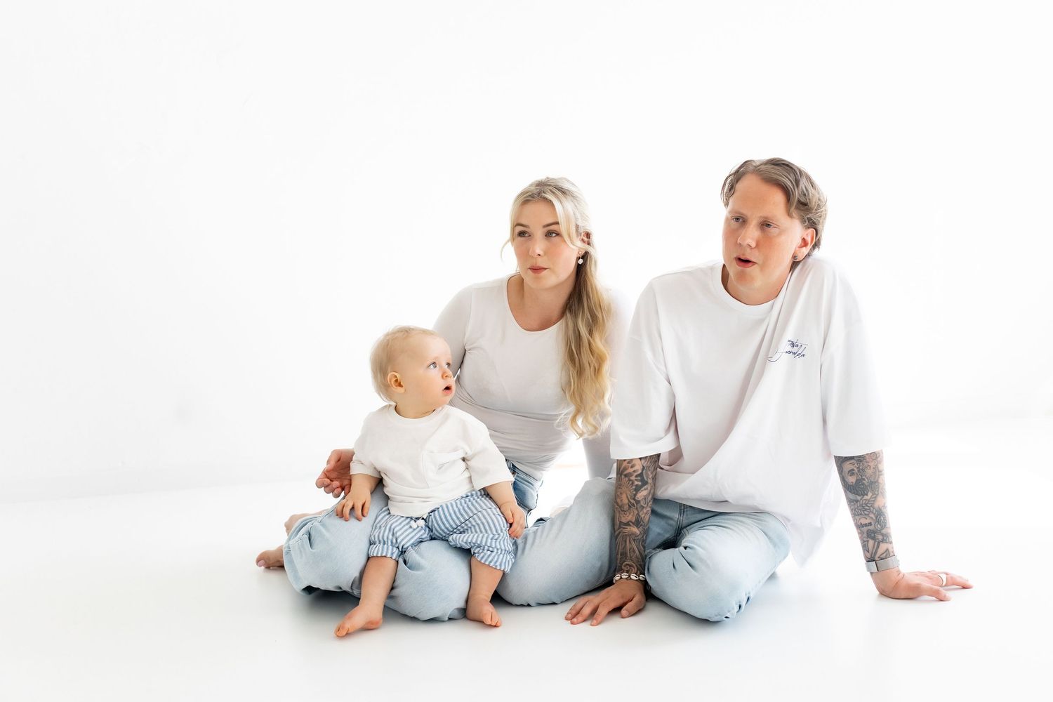 Smiling family of three in white tees and light jeans sitting together on a white studio floor, with a baby between a blonde mother and tatt