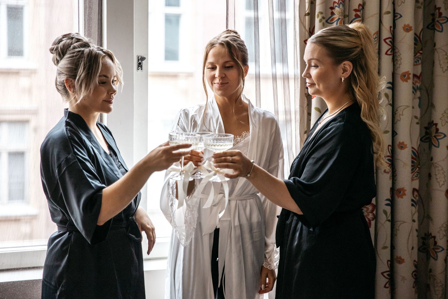 Three women in robes toasting with champagne glasses by a bright window during bridal preparations.