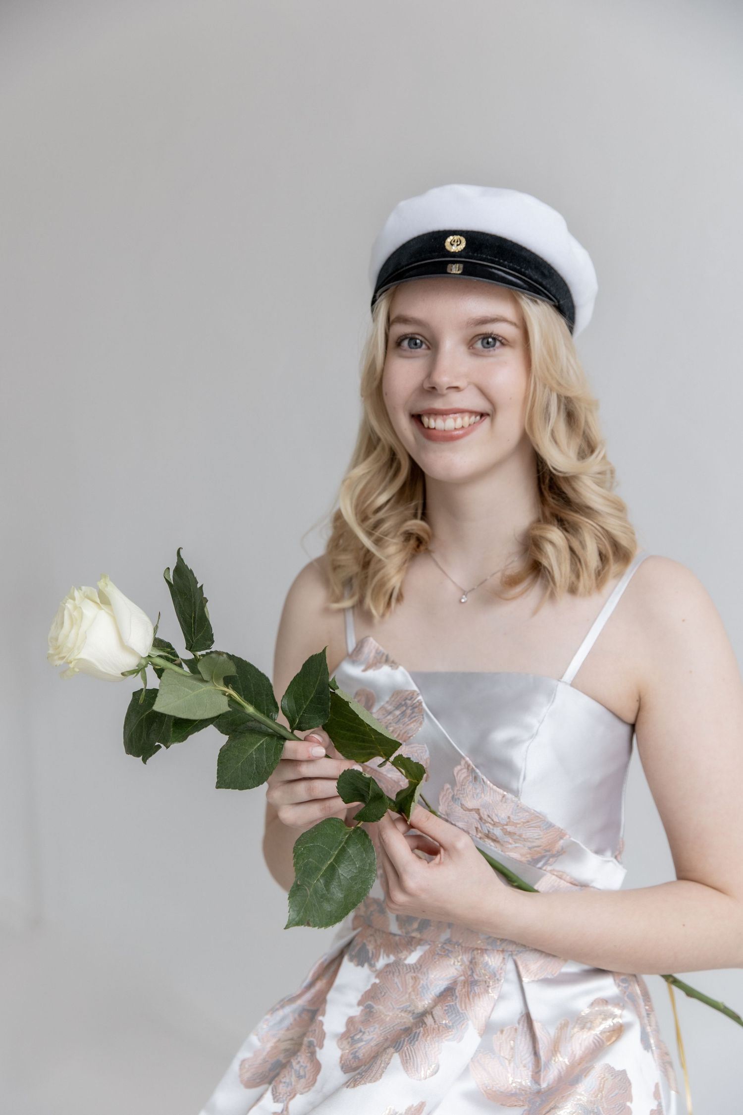 Smiling young woman with wavy blonde hair wears a white student cap and satin floral dress while holding a single white rose.