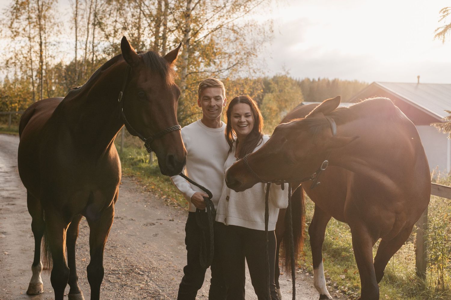 Couple smiling between two brown horses on a sunny rural path surrounded by autumn trees.