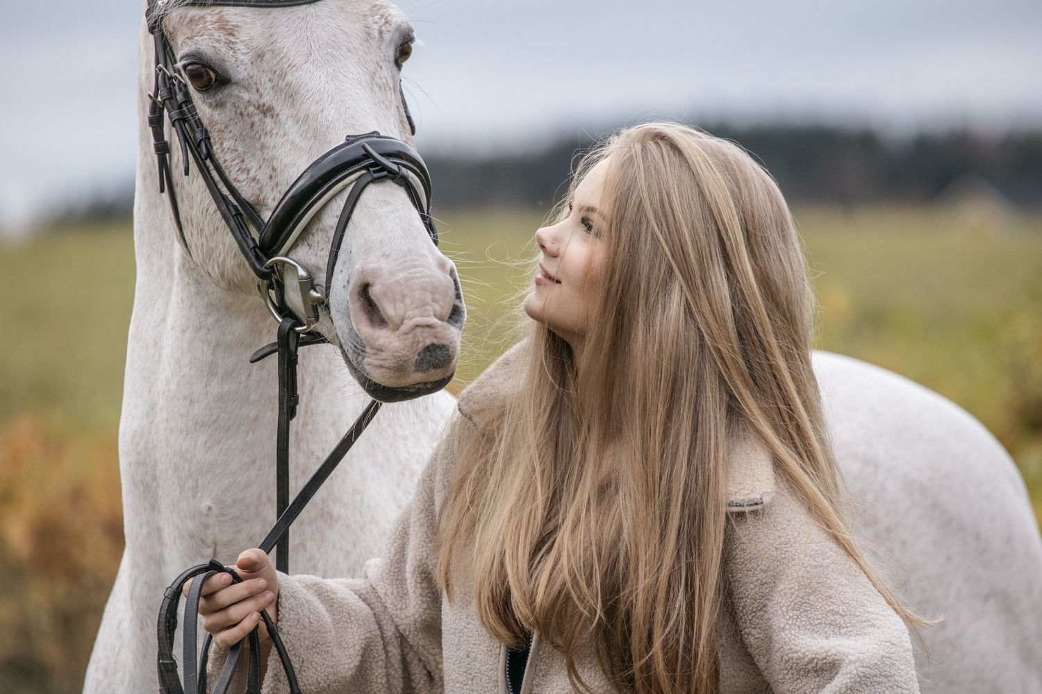 Young blonde woman tenderly bonding with a white horse in a bridle outdoors in a green field.