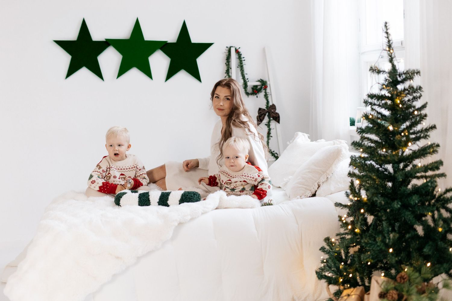 Mother and two babies in matching holiday pajamas sit on a white bed beside a decorated Christmas tree with green star wall decor.
