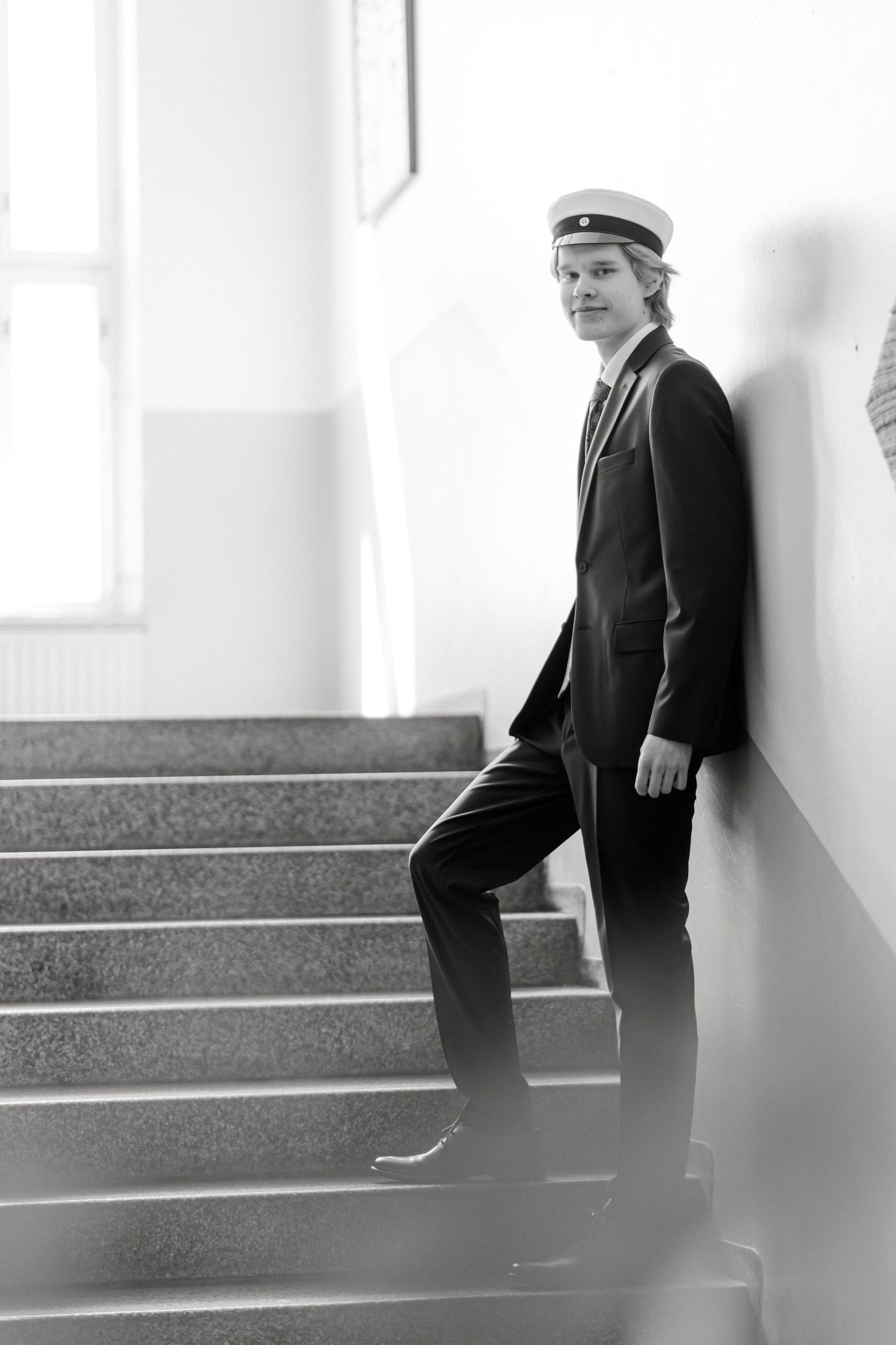 Young man in a dark suit and white student cap leaning against a wall on a stone staircase in a black and white portrait.