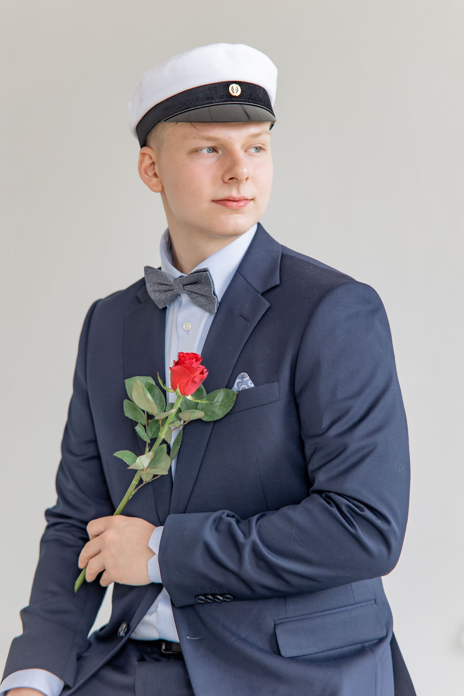 Young man in a navy blazer, gray bow tie and white student cap holds a red rose while looking to the side against a plain light background.
