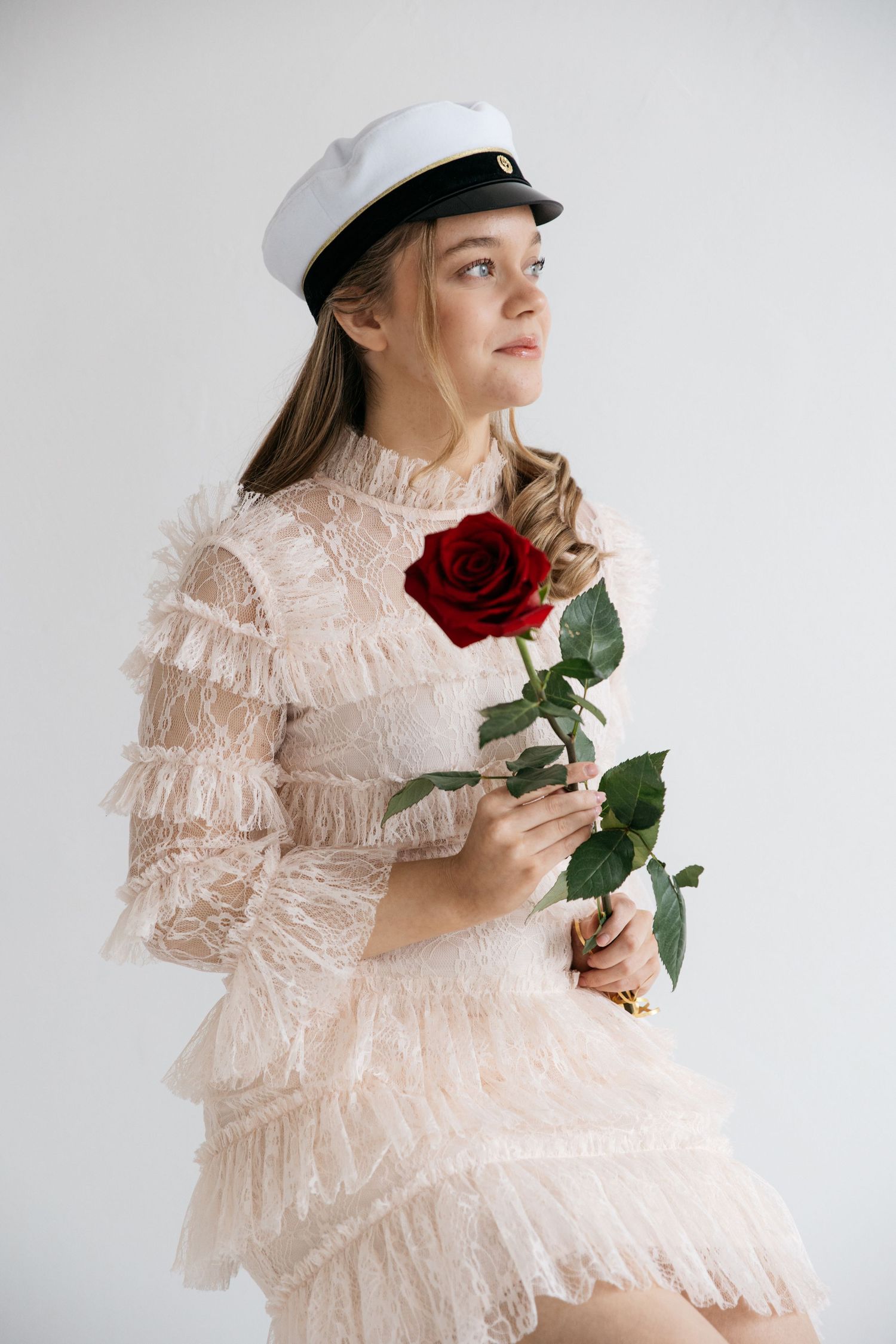 Young woman in a white student cap and blush ruffled lace dress sits holding a red rose against a plain white background.