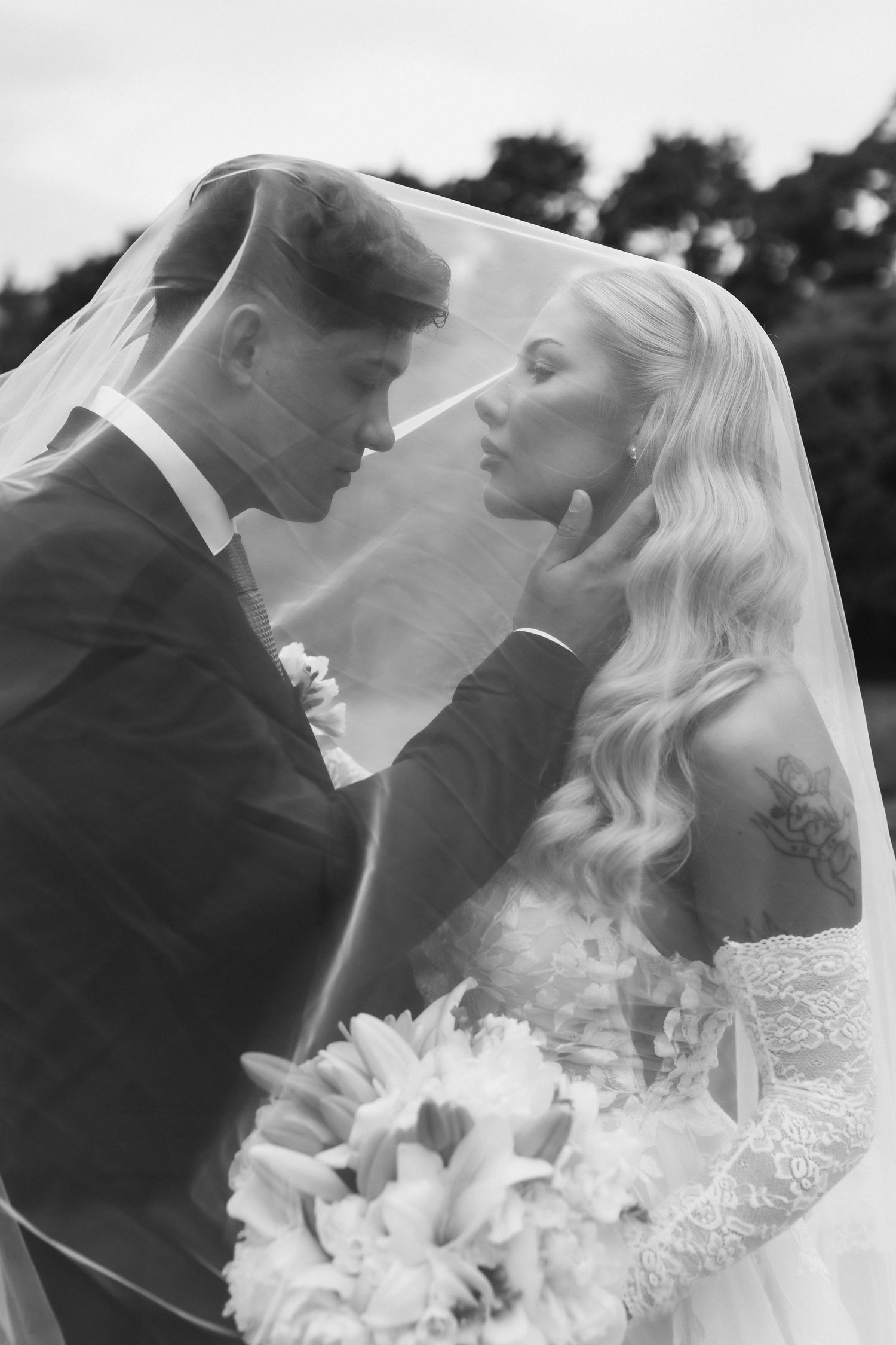 Black and white romantic portrait of bride and groom sharing intimate moment under flowing wedding veil outdoors.