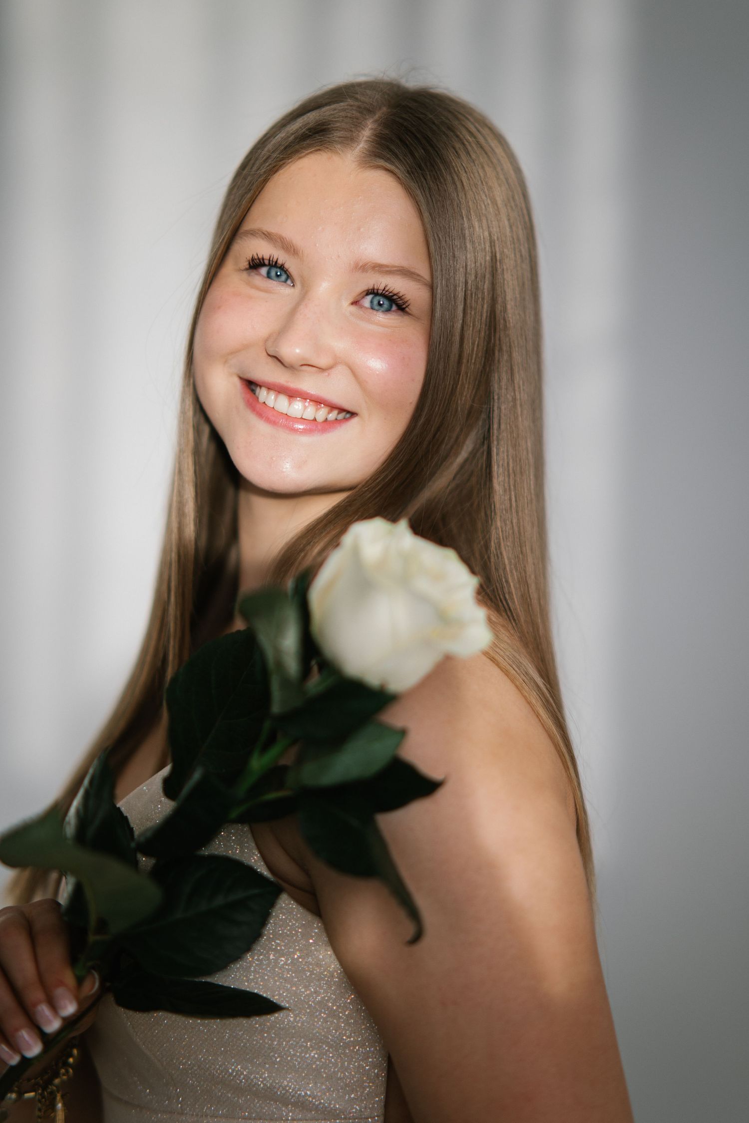 Smiling young woman with long straight hair holding a white rose, wearing a shimmery champagne dress against a soft neutral background.