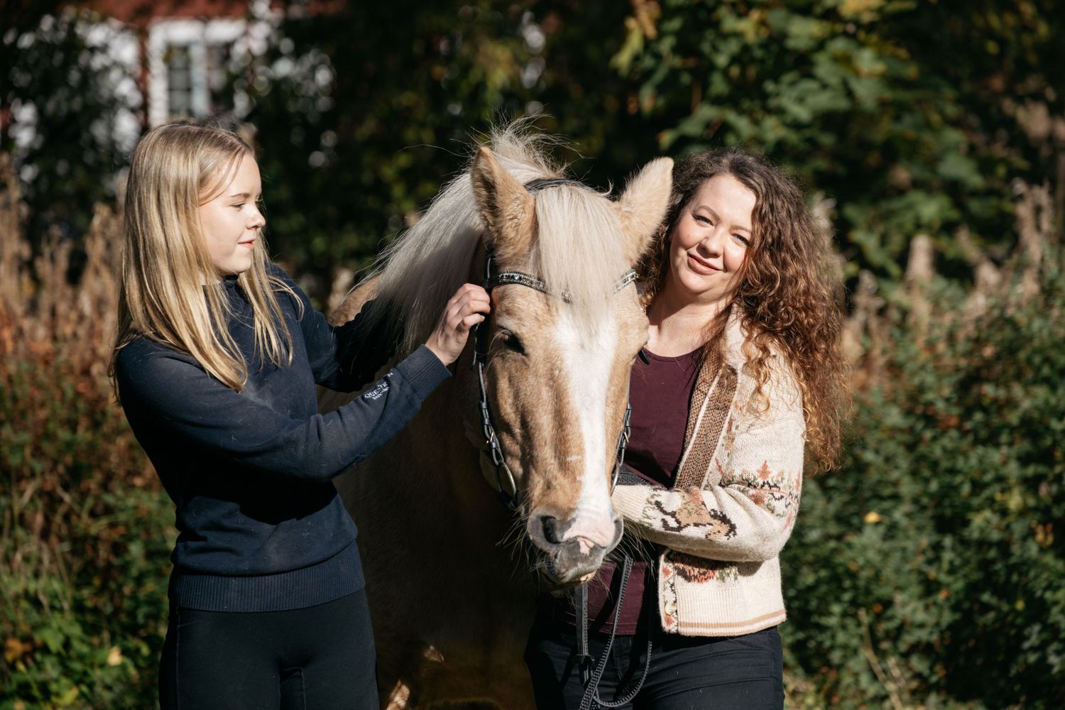 Two women smiling and grooming a white horse outdoors surrounded by autumn trees.