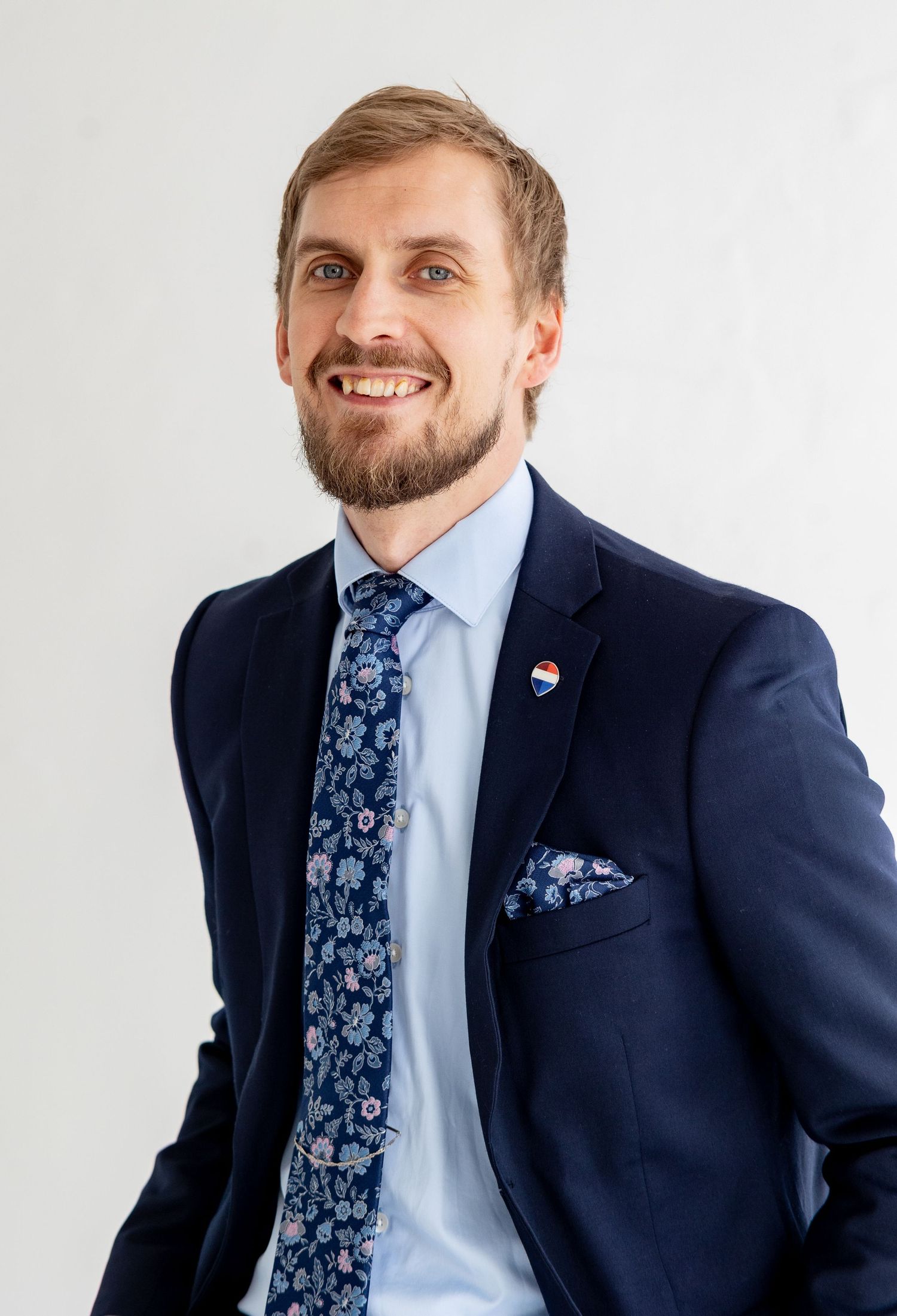 Smiling bearded man in navy floral tie and suit with RE/MAX pin on white background.