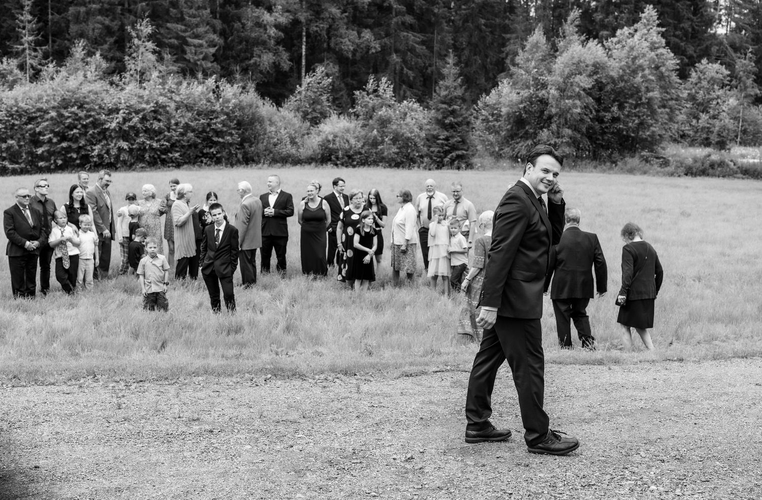 Black and white photo of groom standing alone as wedding guests gather in background outdoors.