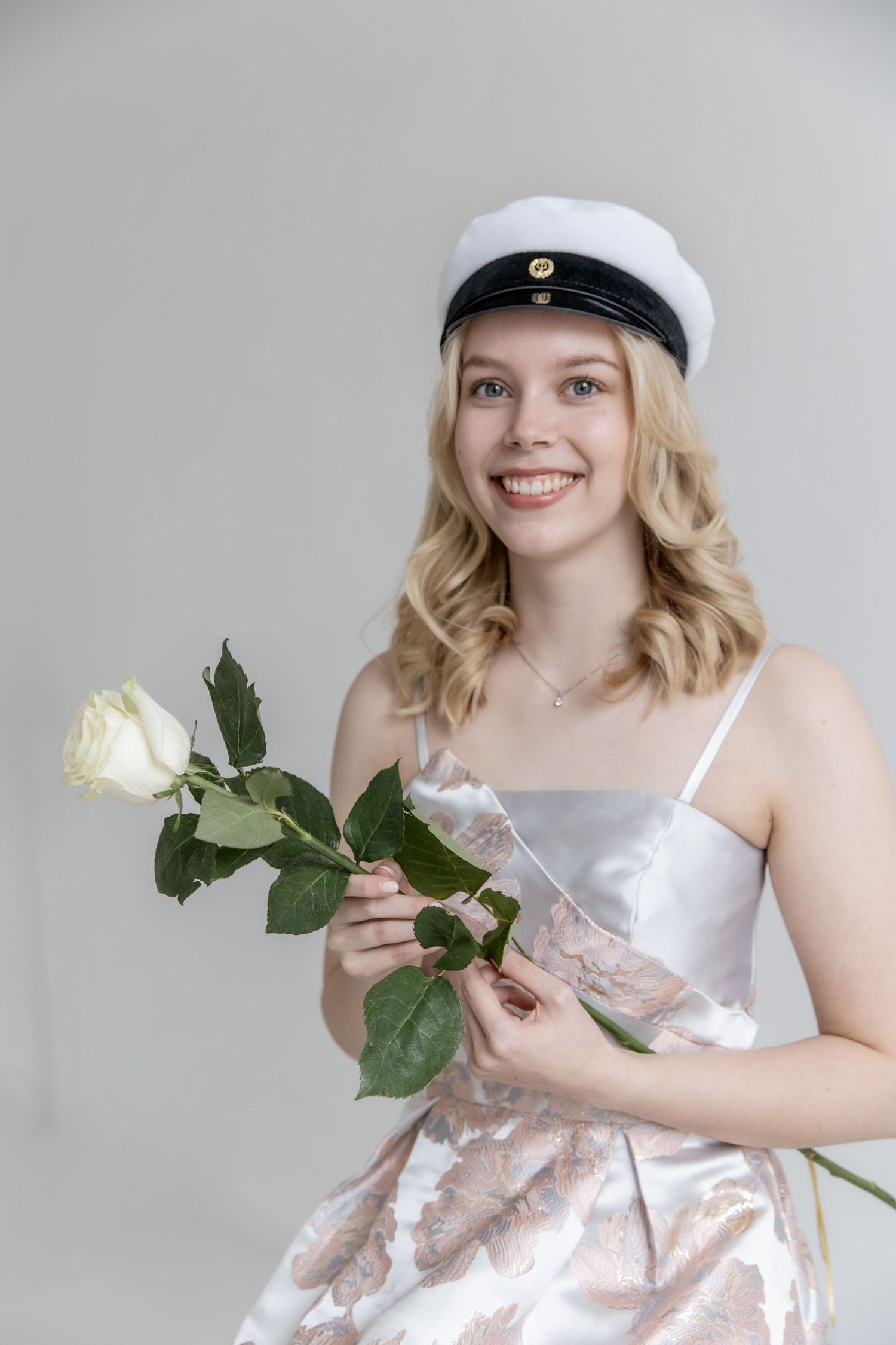 Smiling young woman in a white student cap and silver-pink satin dress holds a white rose against a plain light background.