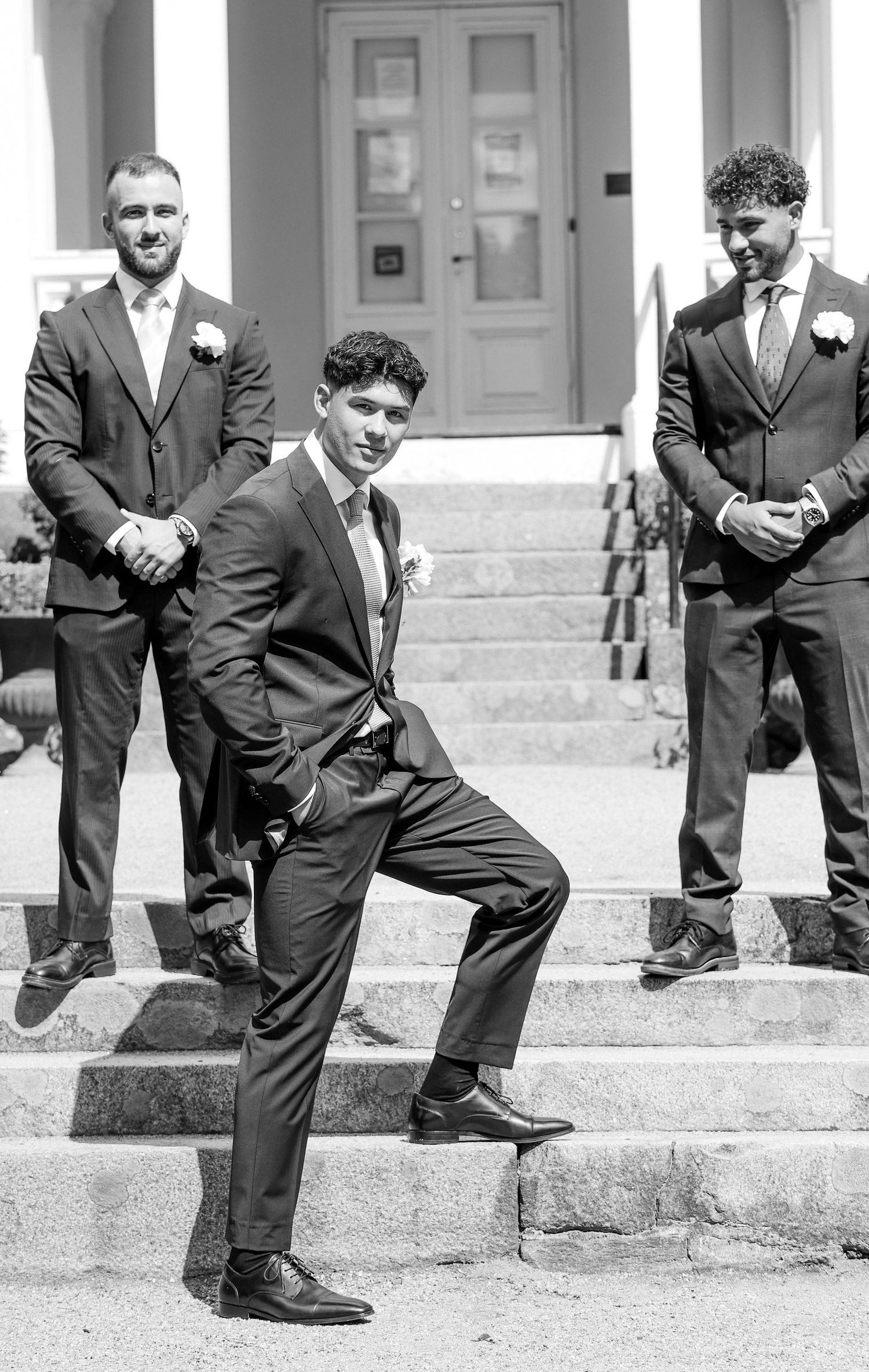 Black and white photo of three groomsmen in dark suits posing on church steps before wedding ceremony.