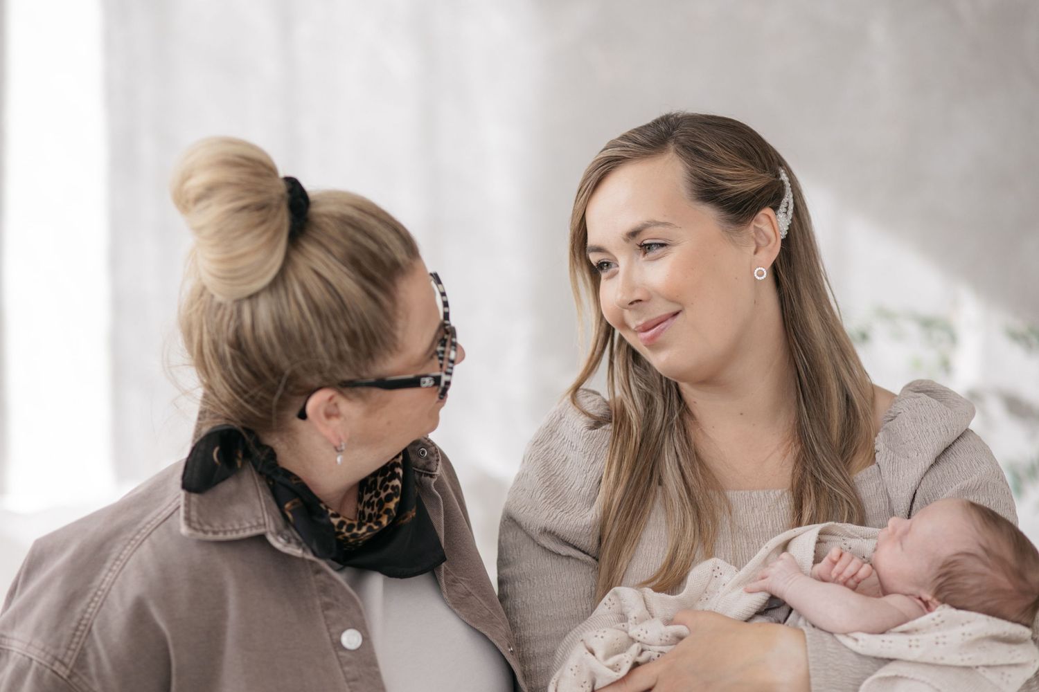Two smiling women in neutral clothing look at each other warmly while one gently holds a sleeping newborn baby wrapped in a soft blanket.