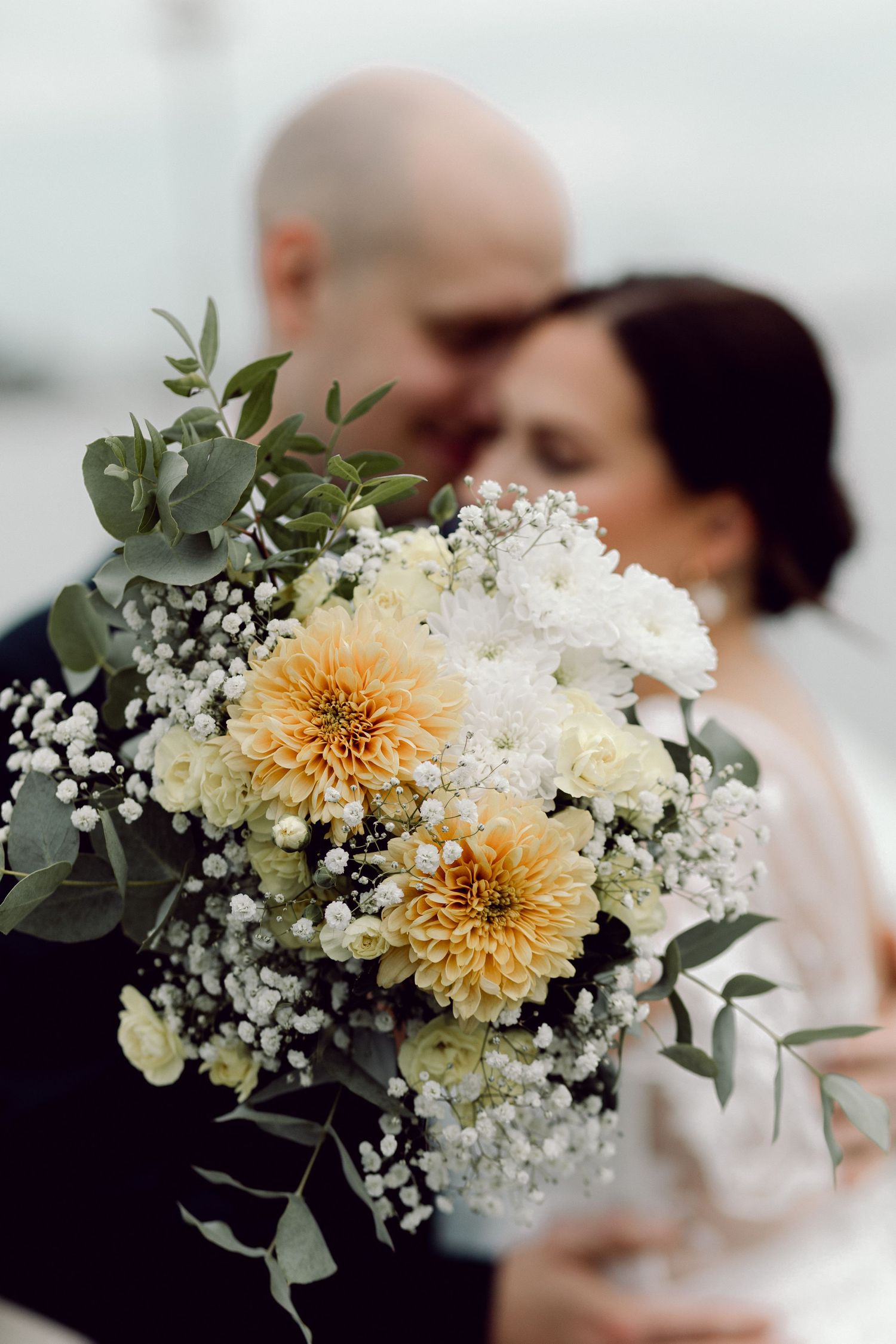 Bridal bouquet of orange dahlias, white roses, and eucalyptus with couple kissing softly in background.