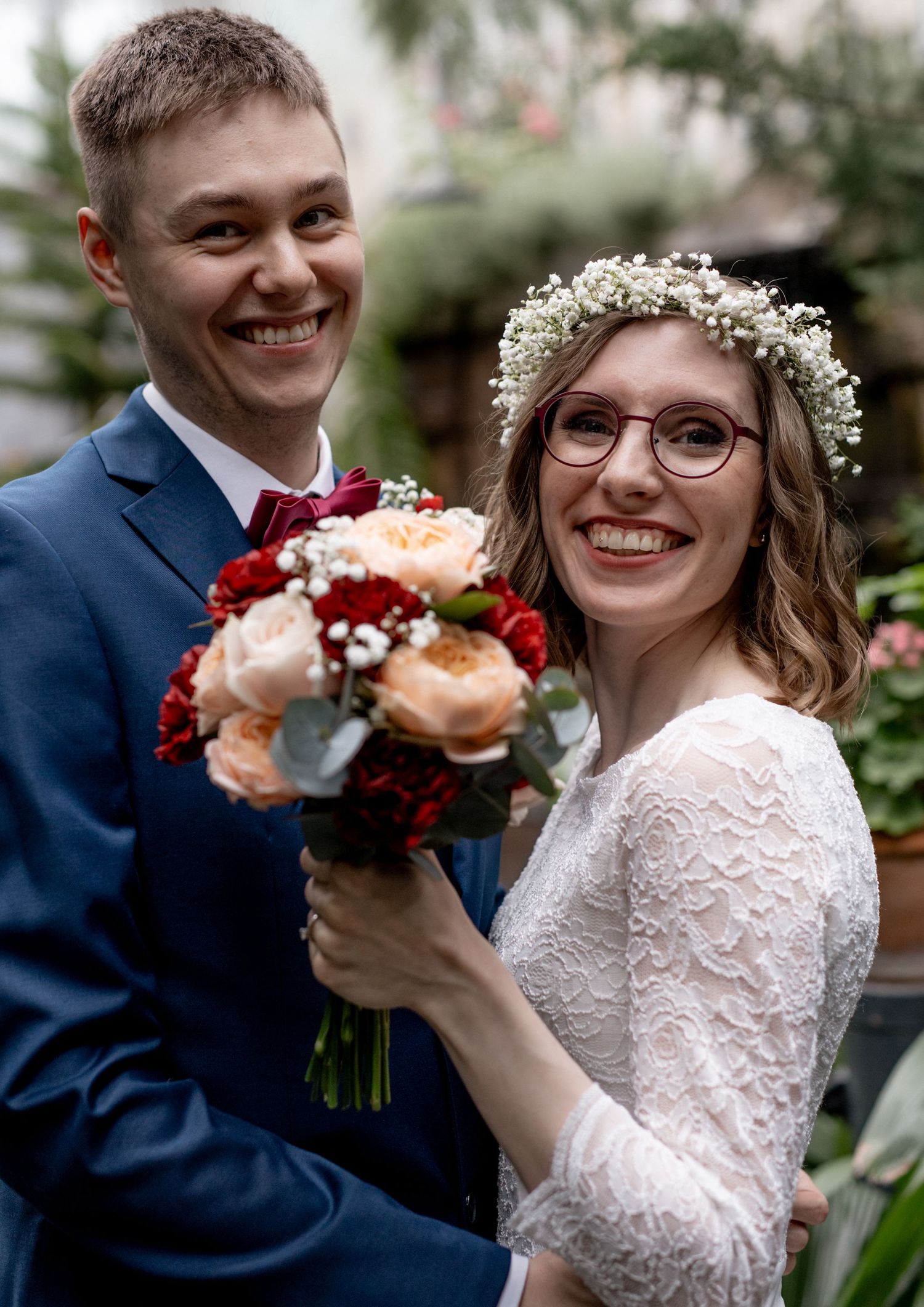 Smiling wedding couple holding red and peach bouquet, bride wearing flower crown and glasses, groom in blue suit.