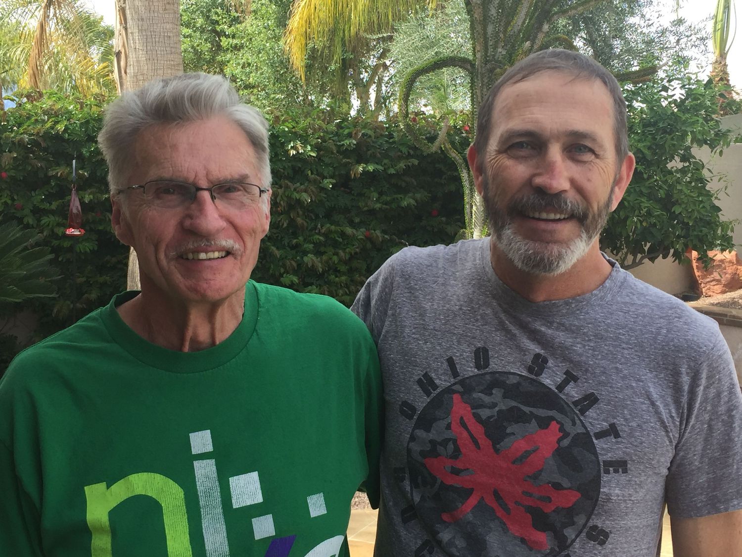 Two smiling men pose outdoors, one in a green shirt and one in a gray graphic tee, with tropical greenery behind them.