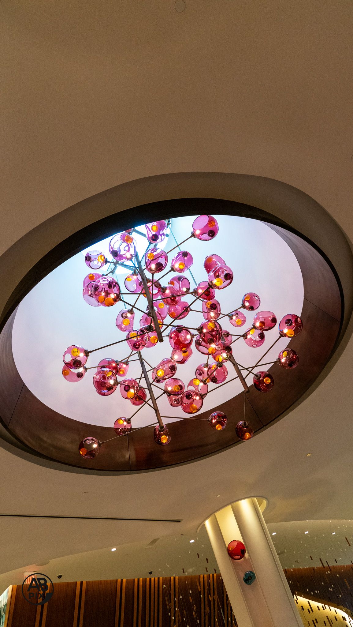 Pink and rose-colored glass ball chandelier suspended within oval ceiling recess in modern lobby.