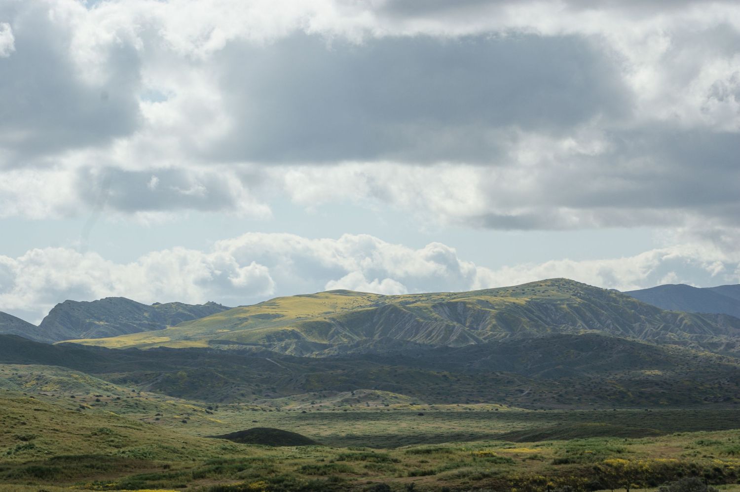 Rolling green hills and rugged mountains under dramatic cloudy sky in a remote wilderness landscape.