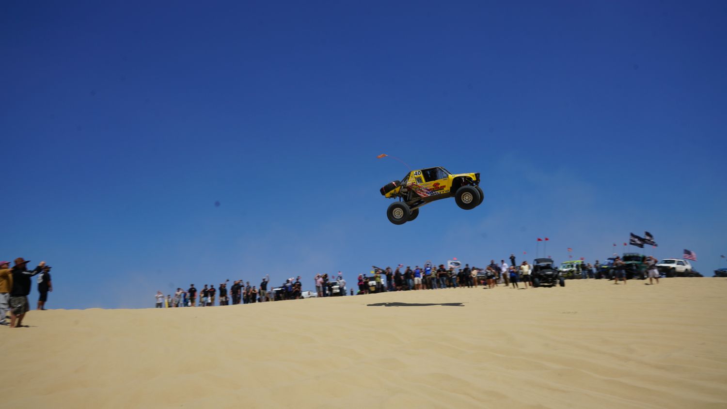 Yellow off-road buggy soaring through blue sky over sand dunes while spectators watch at a desert racing event.
