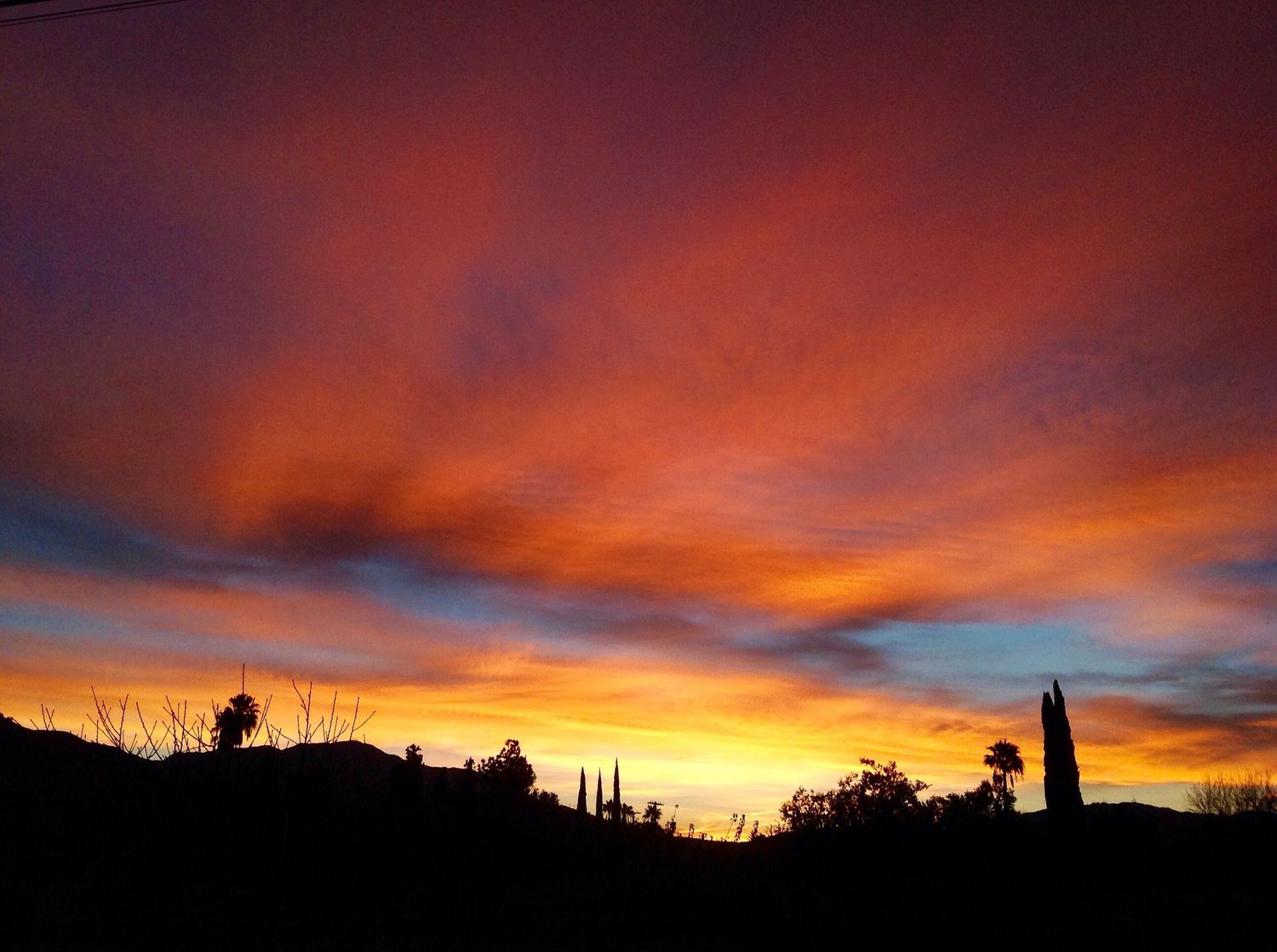 Dramatic fiery orange and red sunset clouds over dark silhouetted hills and trees.