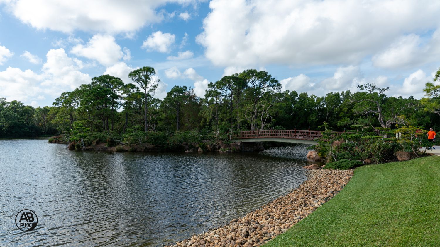 A scenic wooden bridge spans a calm lake surrounded by lush green trees under a partly cloudy sky.