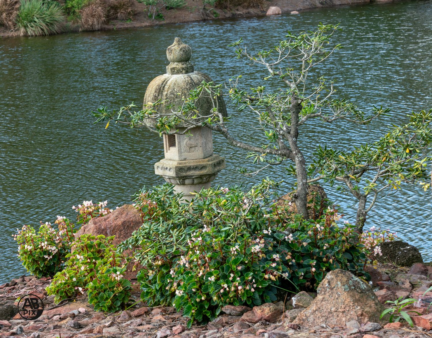 Stone Japanese lantern on a rocky island surrounded by colorful flowers beside a calm pond.