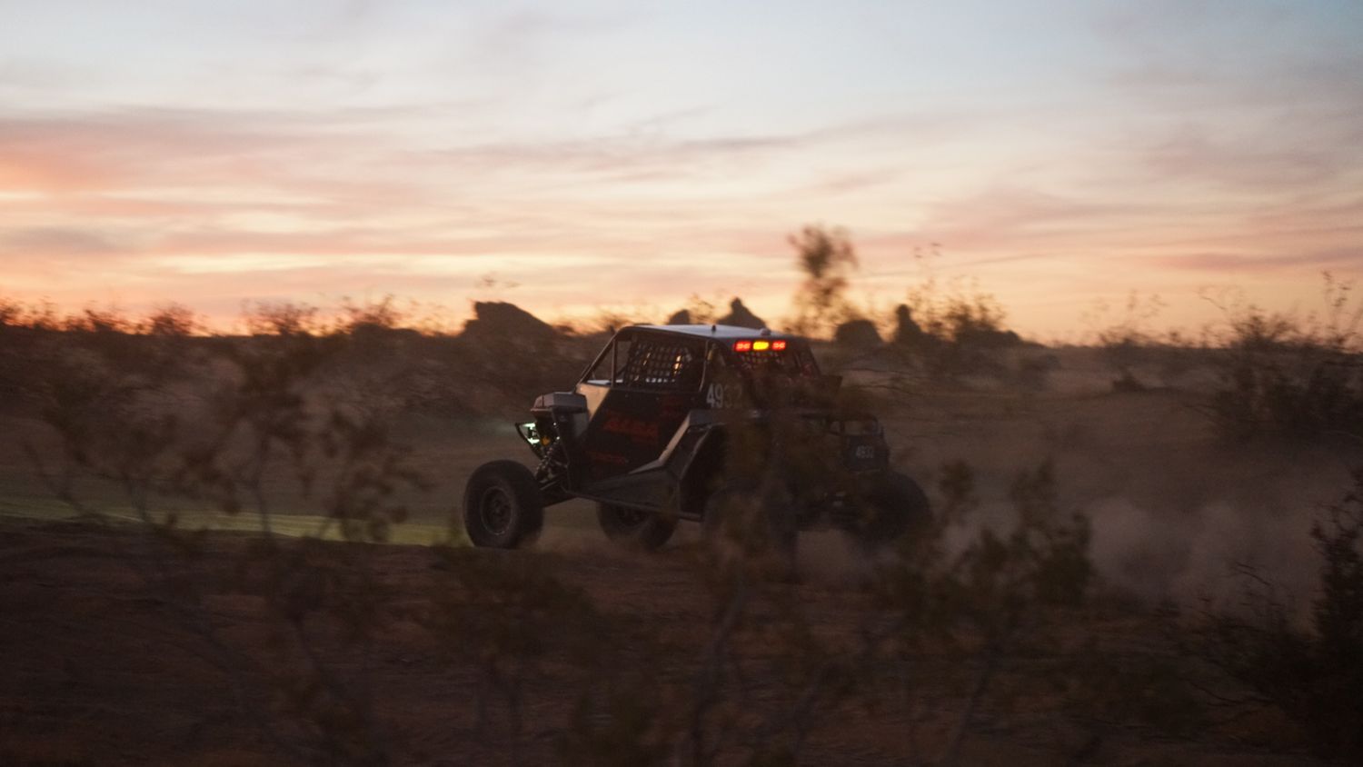 UTV side-by-side vehicle catching air during off-road race at sunset in desert landscape.