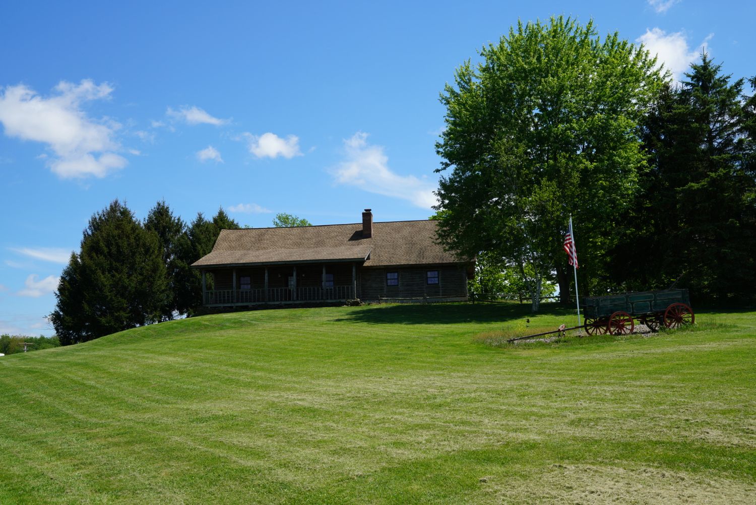 Historic log cabin building on a manicured green hillside with large shade trees and an American flag.