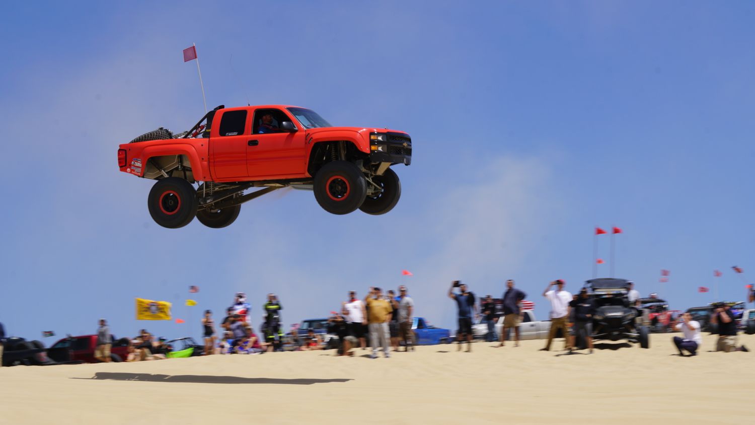 Red off-road trophy truck soars high above sand dunes in front of a large crowd of spectators.