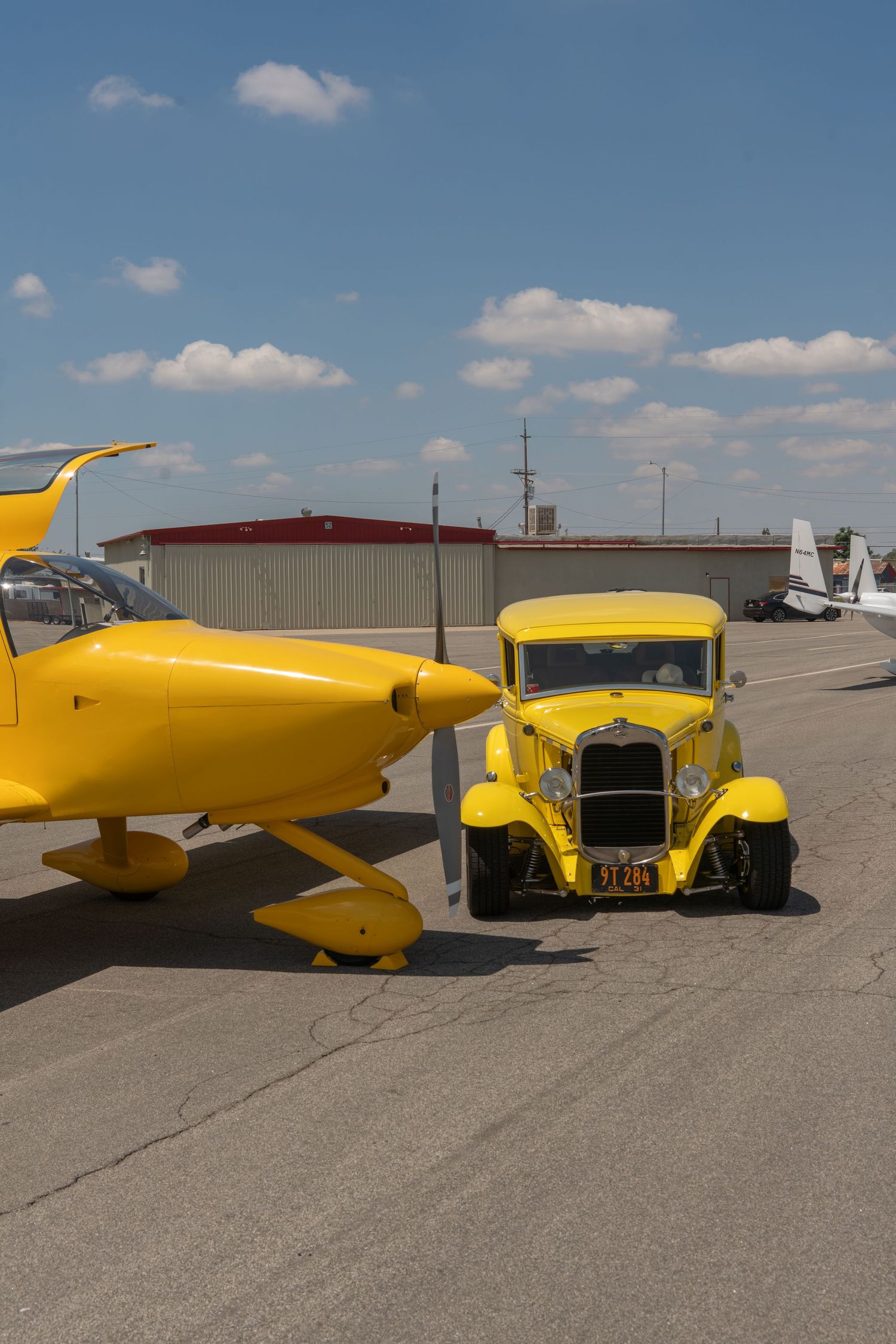 Matching yellow vintage hot rod car parked beside a yellow biplane on an airport tarmac under a blue sky.