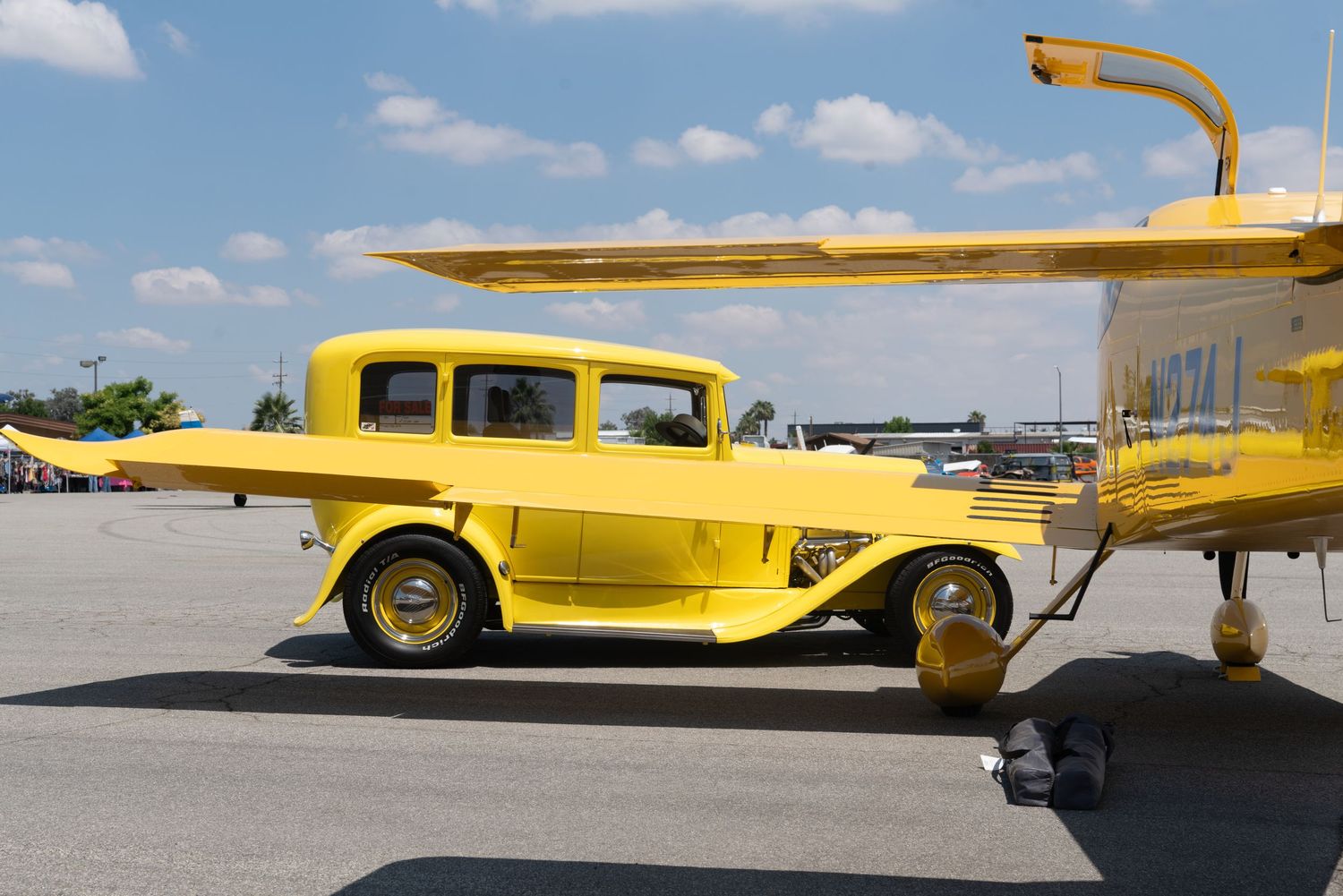 Bright yellow vintage hot rod car parked next to a matching yellow airplane on a sunny airfield tarmac.