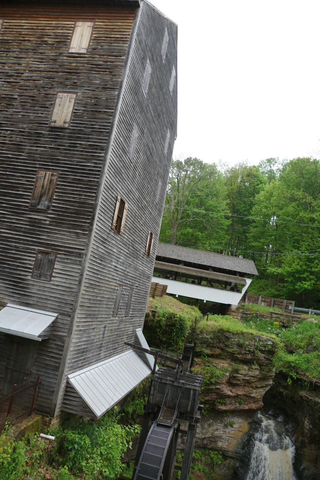 Tall weathered wooden grist mill beside a rocky waterfall surrounded by lush green trees.