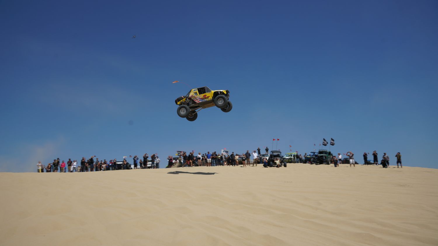 Yellow off-road race buggy launching high into the air over sand dunes before a large crowd of spectators.