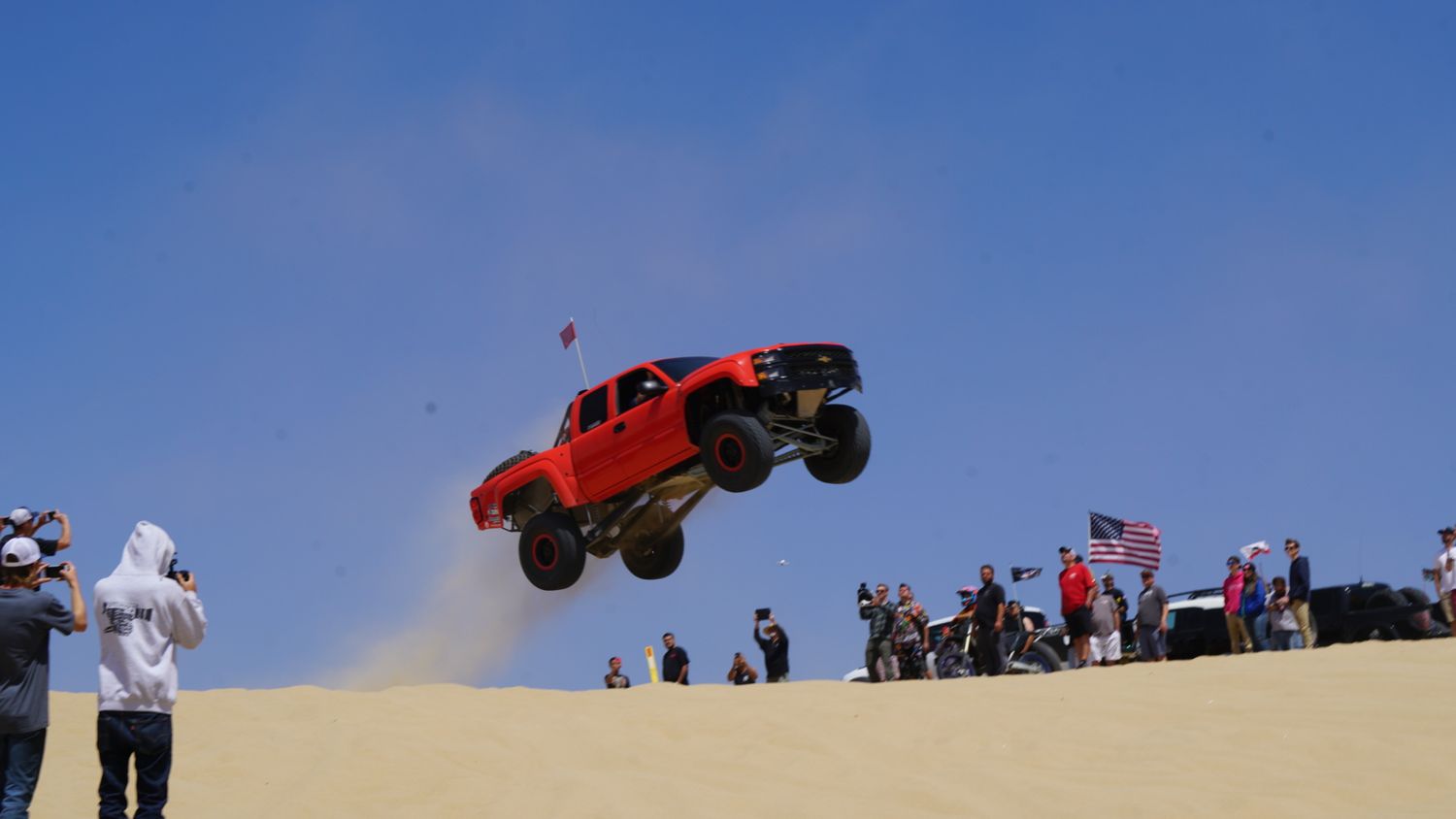 A red off-road truck launching off a sand dune in front of spectators at an outdoor racing event.