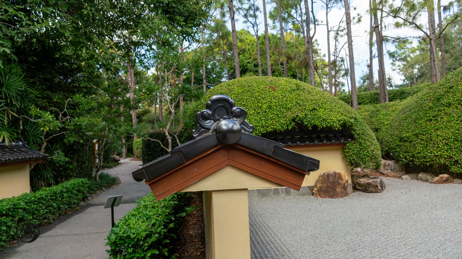 Traditional Japanese gate with ornate black roof tile and decorative finial surrounded by manicured green hedges.