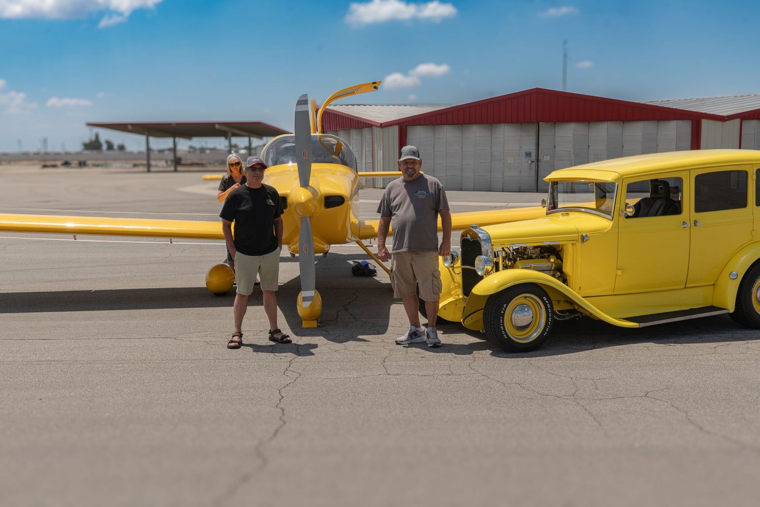 Two men pose beside a matching yellow vintage hot rod car and small yellow airplane on a sunny airport tarmac.