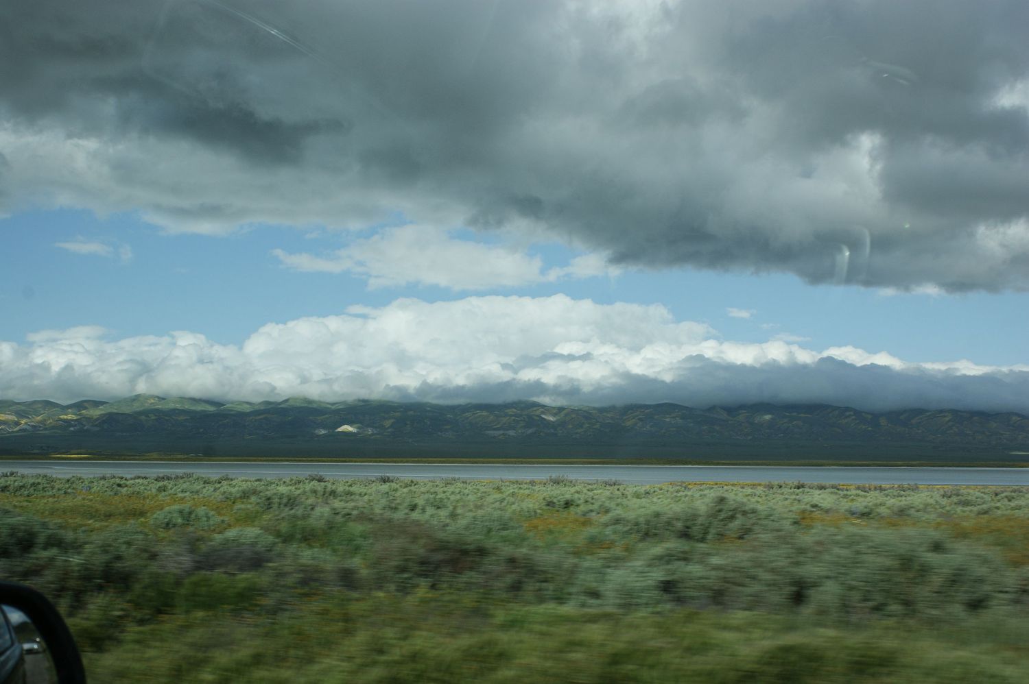 Scenic lake view with snow-capped mountains under dramatic storm clouds from car window.