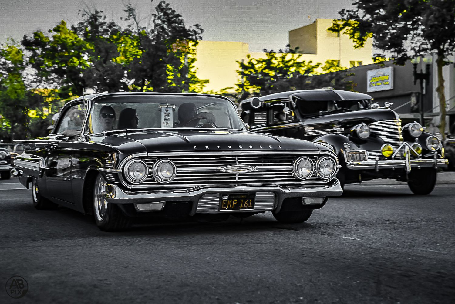 Vintage black Chevrolet Impala lowrider cruising on a street at a classic car show at dusk.