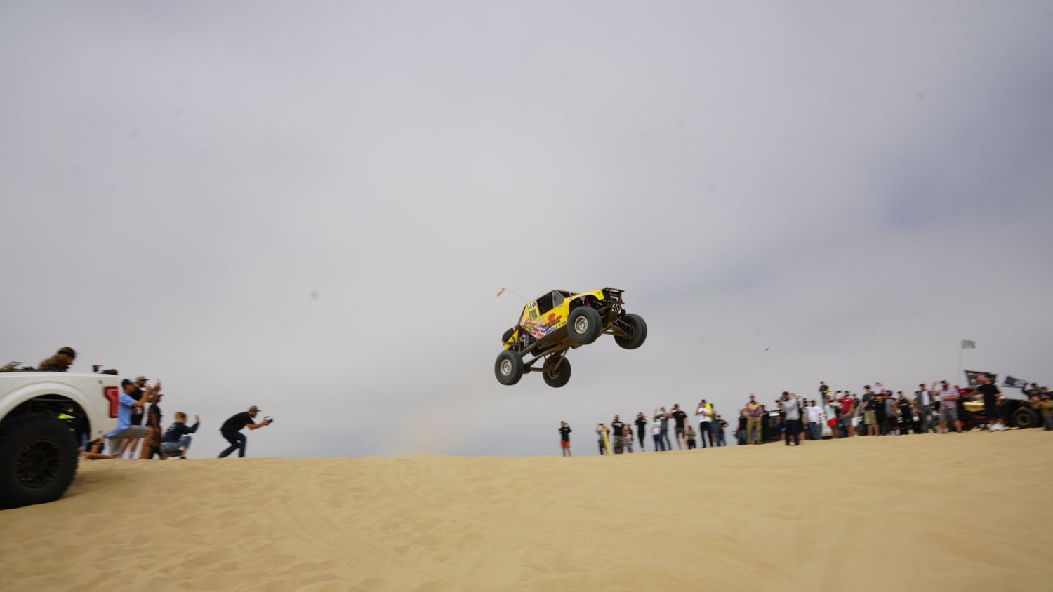Yellow dune buggy soaring through overcast sky over sandy dunes as excited spectators watch the stunt.