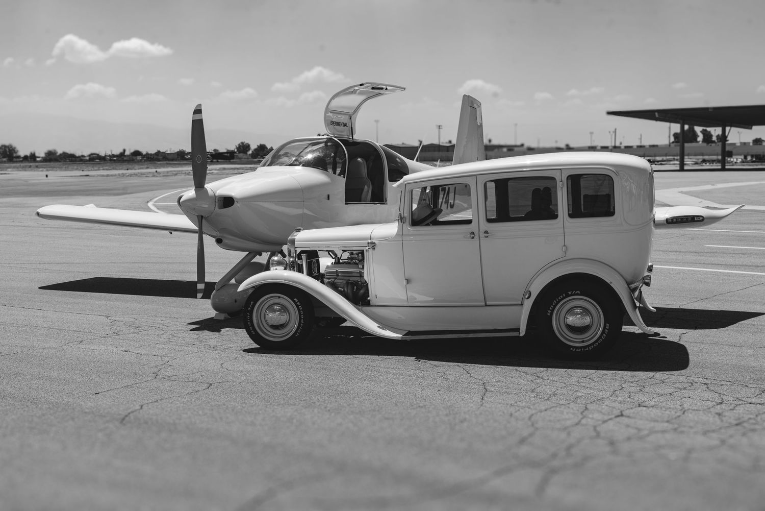 Vintage black and white photo of a classic car parked beside a propeller airplane on an airfield tarmac.