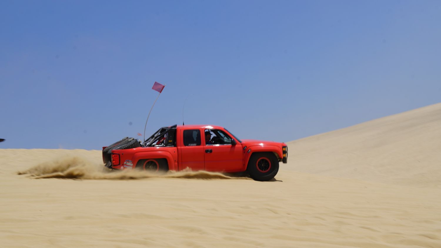 Red off-road trophy truck kicks up sand while racing across vast open desert dunes under blue sky.