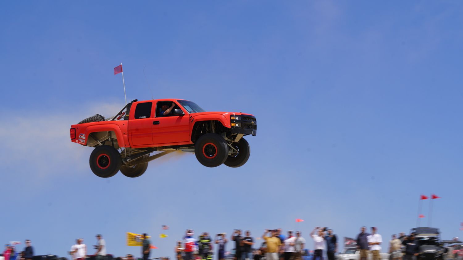 A red off-road trophy truck soaring high in the air against a blue sky at a sand dune event.