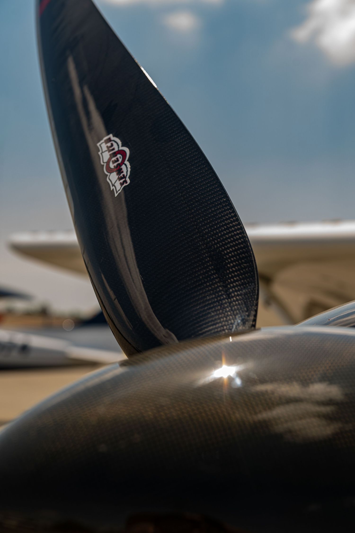 Close-up of a carbon fiber aircraft propeller blade against a blue sky with sunlight glare.