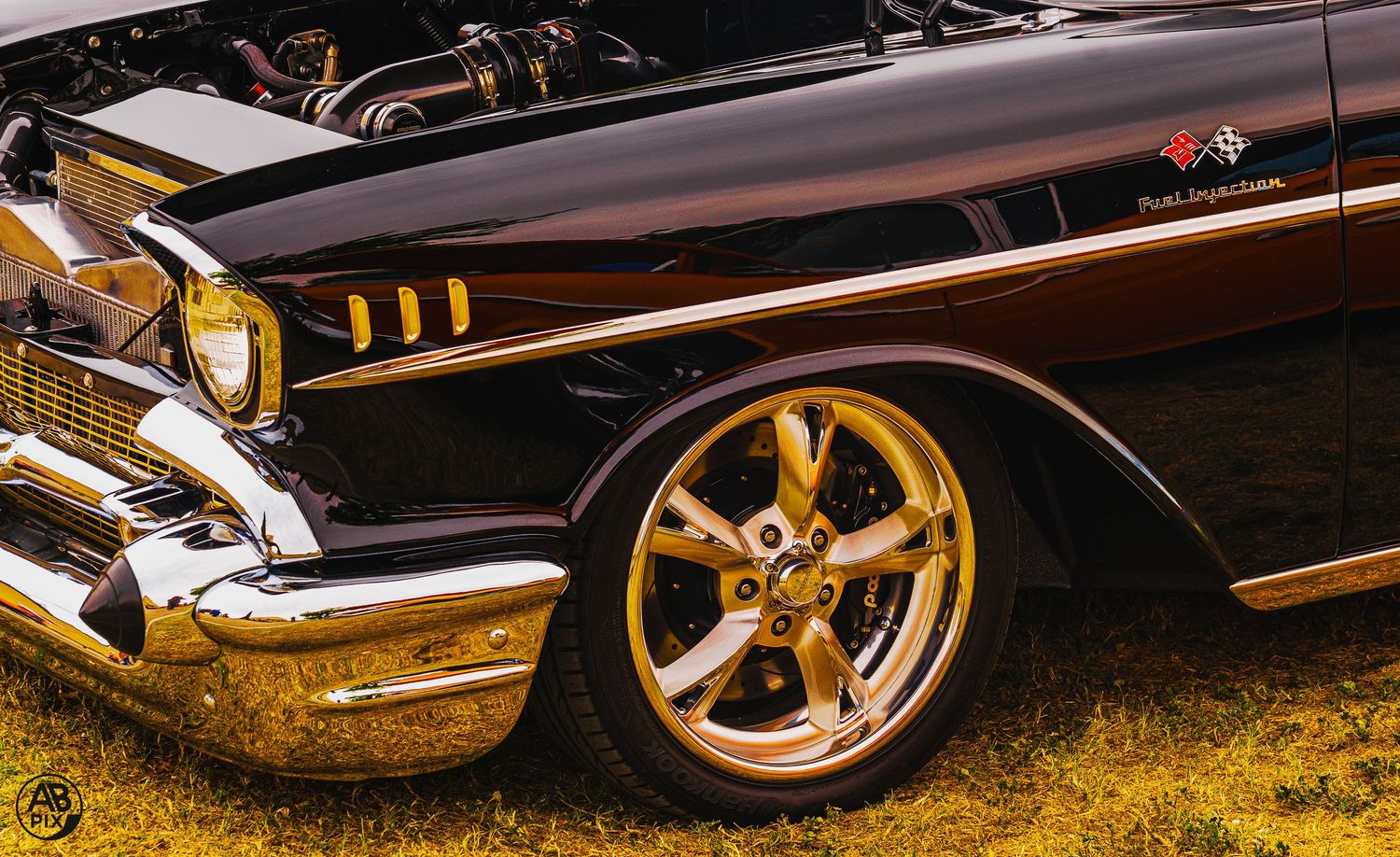 Side detail of a customized black 1957 Chevy with gold alloy wheels on green grass.