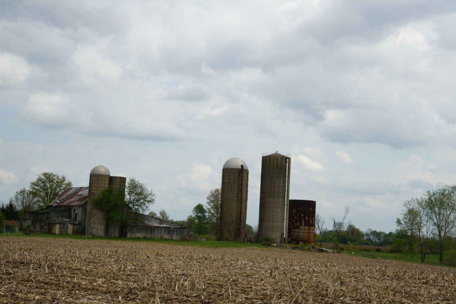 Abandoned farm with weathered barn and three silos visible across a plowed field under a cloudy sky.